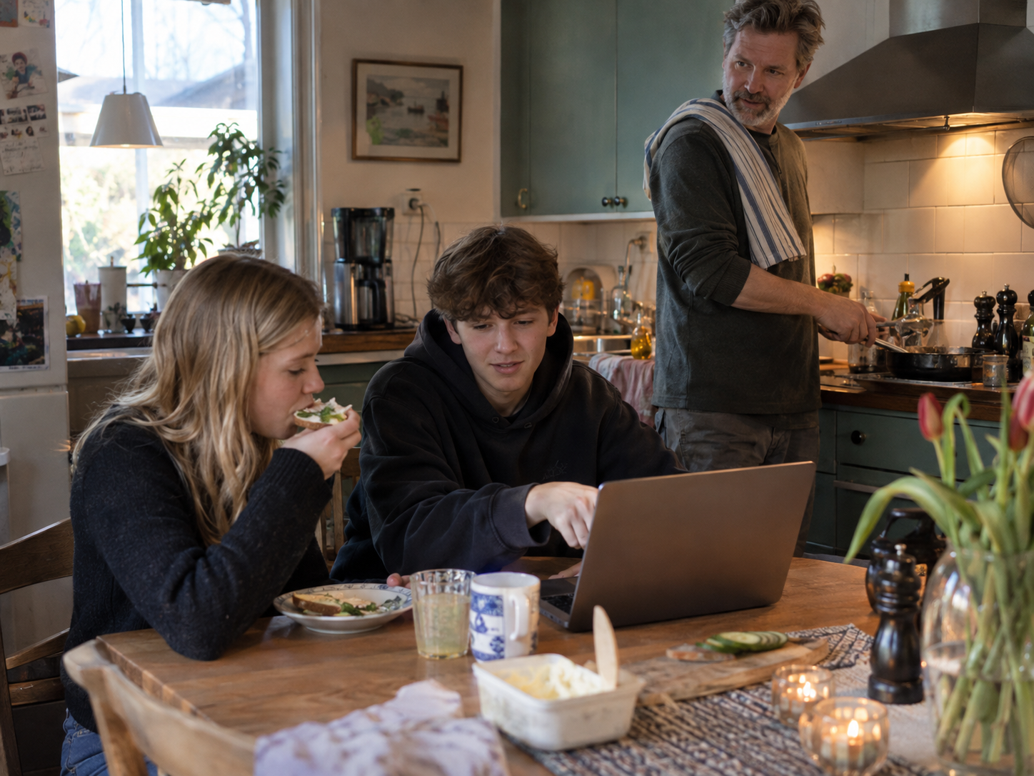 En familj i köket: två ungdomar vid en laptop vid köksbordet medan en vuxen lagar mat.
